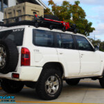 Rear right view of a Nissan GU Patrol Wagon in White fitted with a Airbag Man Coil Air Kit 3" Inch Lift Kit & Superior Tie Rod Comp Spec 4340m Solid Bar whilst on the hoist at Superior
